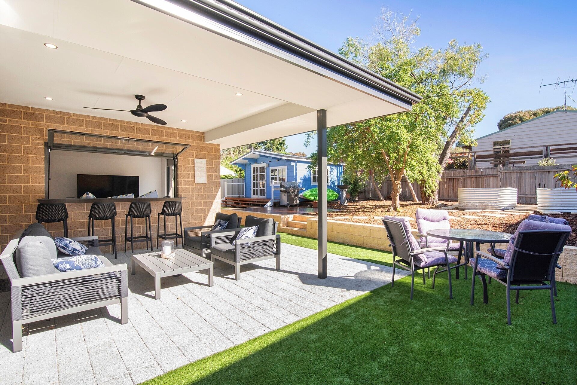 An outdoor patio with a brick wall, a ceiling fan, and a TV mounted on the wall. There are multiple seating areas with chairs and a table.