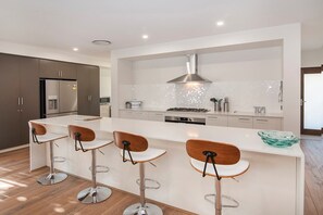 A modern kitchen with a central island, bar stools, stainless steel appliances, and a backsplash. - The Milly (Dunsborough)