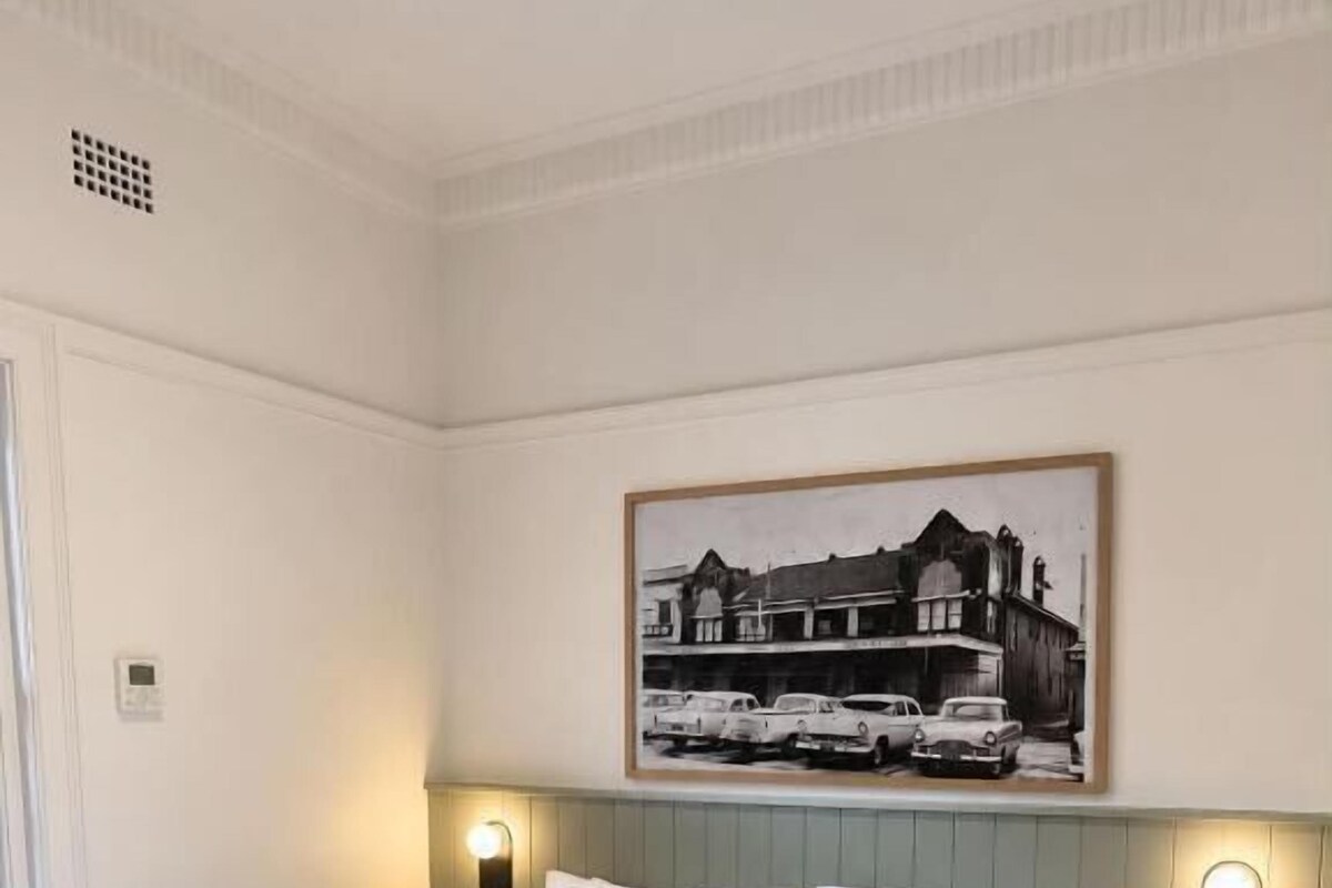 Quarto de hotel com uma cama grande, duas mesas de cabeceira, luminária fixada na parede e uma moldura com imagem sobre a cama.