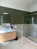 Un baño con lavamanos blanco, tocador de madera, un espejo grande, paredes con azulejos verdes y una cabina de ducha de vidrio.