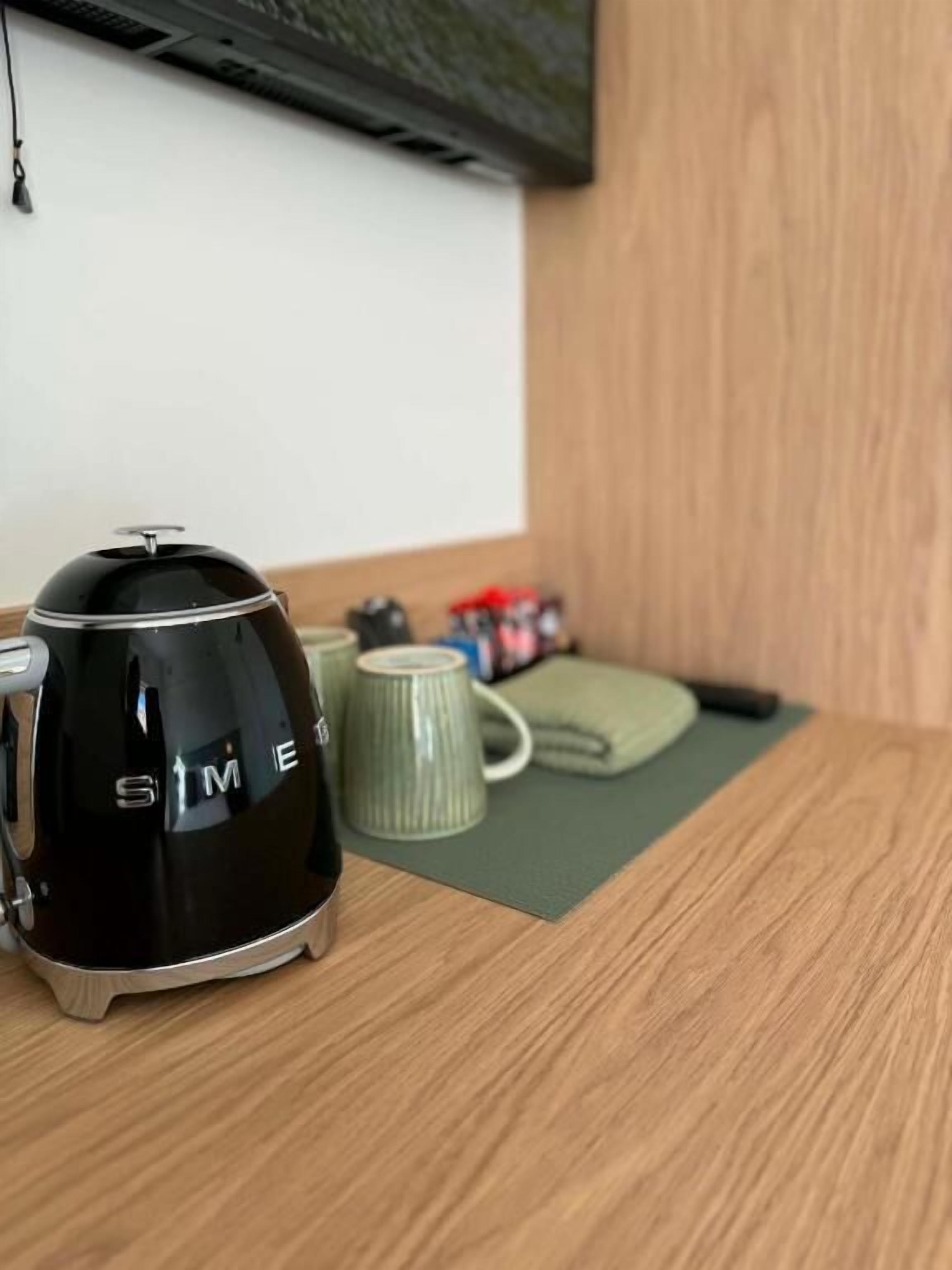 A black coffee maker on a wooden table with a green mat, a white mug, and a green cushion.