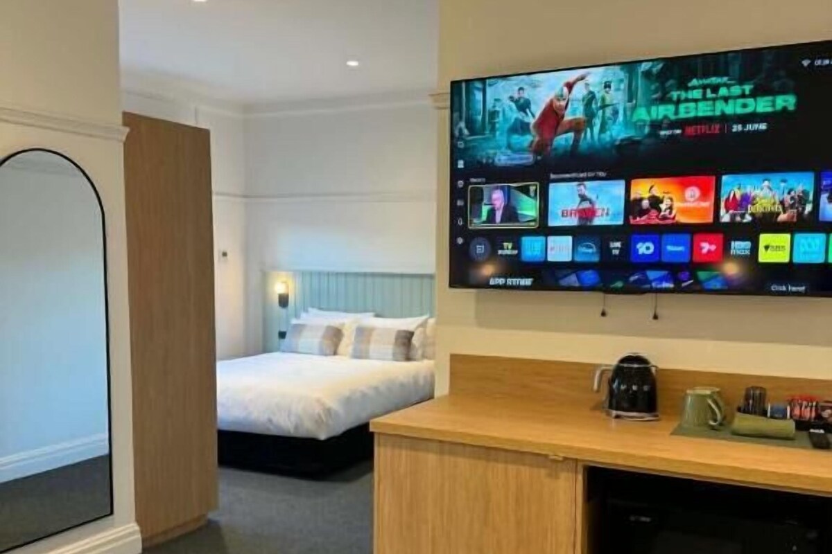 Quarto de hotel com uma TV de tela plana grande fixada na parede, exibindo vários canais e um pôster de filme. Há uma cama com cabeceira branca e uma mesa de cabeceira com uma cafeteira.