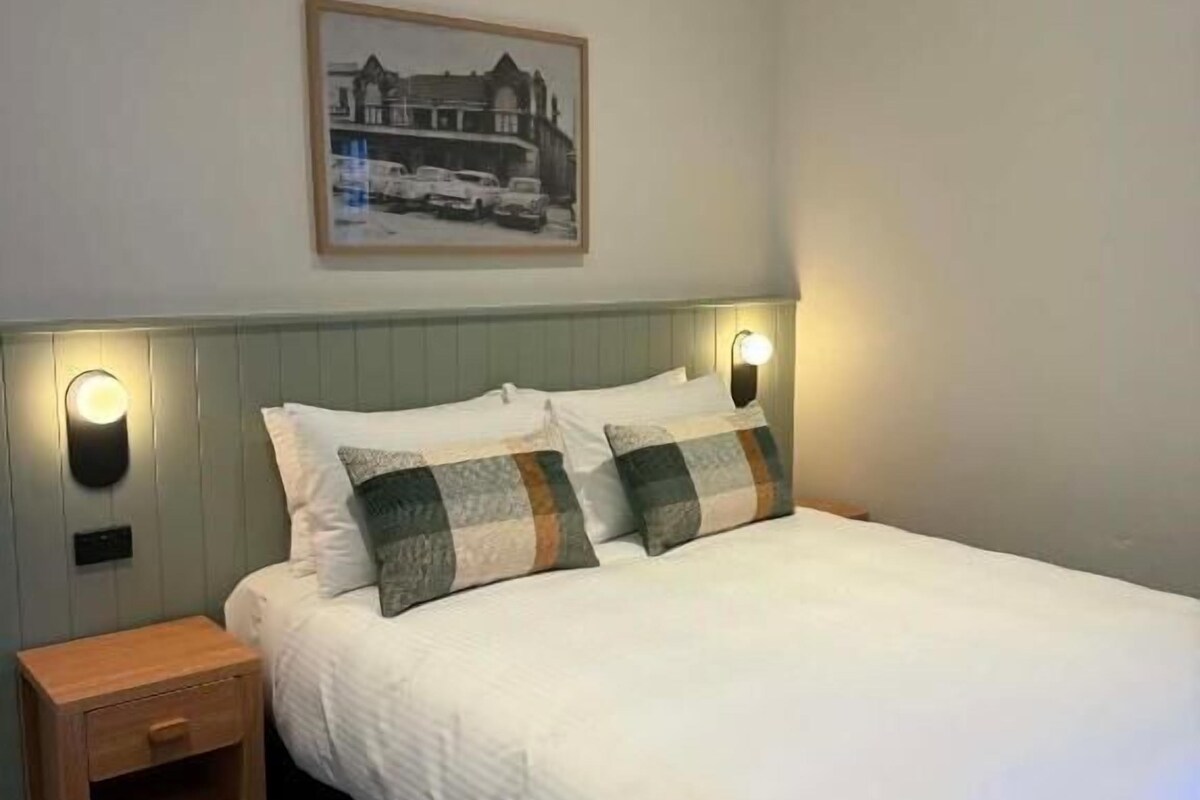 Cama bem arrumada com cabeceira, mesa de cabeceira e luminária fixada na parede.