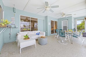 Ein Zimmer mit einem weißen Sofa, einem Esstisch mit Stühlen, einem Deckenventilator und Blick durch eine Glastür.