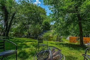A grassy outdoor area with a metal bench and a wicker chair, surrounded by trees and a wooden fence. - Lucky's Cottage | Hot Tub+Dog Friendly+Fire Pit (Rossville)