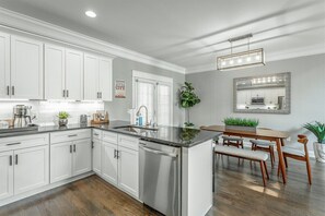 A modern kitchen with white cabinetry, stainless steel appliances, and a dining area with wooden furniture. - The Place | Luxe 3BR | Ideal Location Close to DT! (Chattanooga)