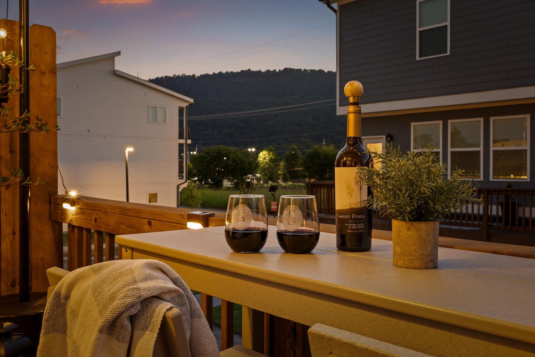 A table with a bottle of wine, two glasses, and a potted plant on a balcony overlooking a residential area.