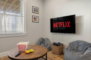 Ein gemütliches Wohnzimmer mit einem runden Holztisch, einer Popcornschüssel, einer Fernbedienung und zwei Sitzsäcken. Ein an der Wand montierter Fernseher zeigt das Netflix-Logo.