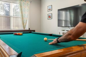 Ein Billardtisch mit Kugeln, ein Fenster mit Jalousien und ein Fernseher an der Wand.