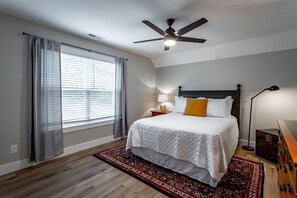Ein Schlafzimmer mit einem Bett, einem Deckenventilator, einem Nachttisch mit Lampe, einem Fenster mit Vorhängen und einem gemusterten Teppich.
