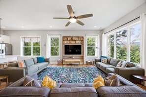 Un salon spacieux doté d’un grand téléviseur à écran plat, d’un ventilateur de plafond et offrant une vue sur l’extérieur grâce à de grandes fenêtres.
