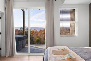 A bedroom with a large sliding glass door leading to a balcony, a bed with a white comforter and towels, and a window with blinds. - Urban Vista | A-Frame | Theatre + Pet Baby Goats! (Rossville)