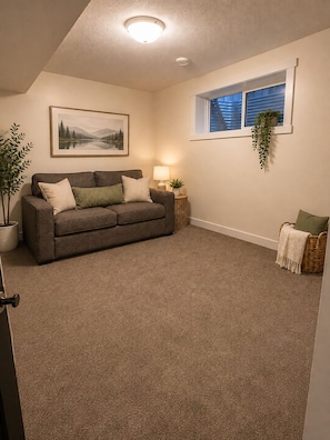 A living room with a gray sofa, a framed landscape painting, a side table with a lamp, a hanging plant, and a window with blinds. - Family-Friendly 5BR Home| Basement Included (Edmonton)