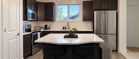A modern kitchen with dark cabinetry, stainless steel appliances, a central island with stools, and a window providing natural light.