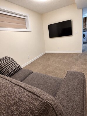 Ein Wohnzimmer mit einem an der Wand befestigten Flachbildfernseher, einem grauen Sofa und einem Fenster mit Jalousien.