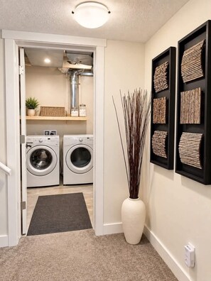 A laundry room with two washing machines, a small shelf with a plant and jars, and a large vase with tall reeds. - Family-Friendly 5BR Home| Basement Included (Edmonton)
