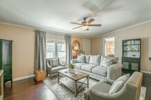 Uma sala de estar com sofá, cadeiras, mesa de centro, ventilador de teto e janela com cortinas.