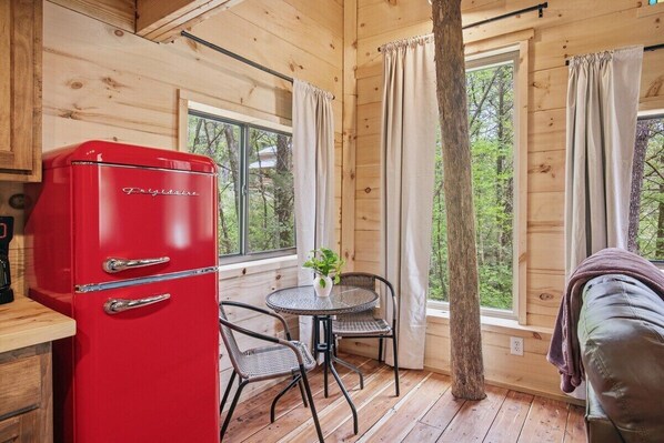 A cabin interior with a red refrigerator, a round table with chairs, and a window with curtains. - Cedarbrook Treehouse | Rustic Escape • Hot Tub! (Trenton)