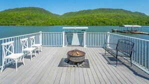 Eine Holzterrasse mit Feuerstelle, weißen Stühlen und einer Bank mit Blick auf einen See und Hügeln im Hintergrund.
