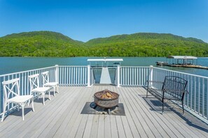 Um deck de madeira com lareira, cadeiras brancas e um banco, com vista para um lago e colinas ao fundo.