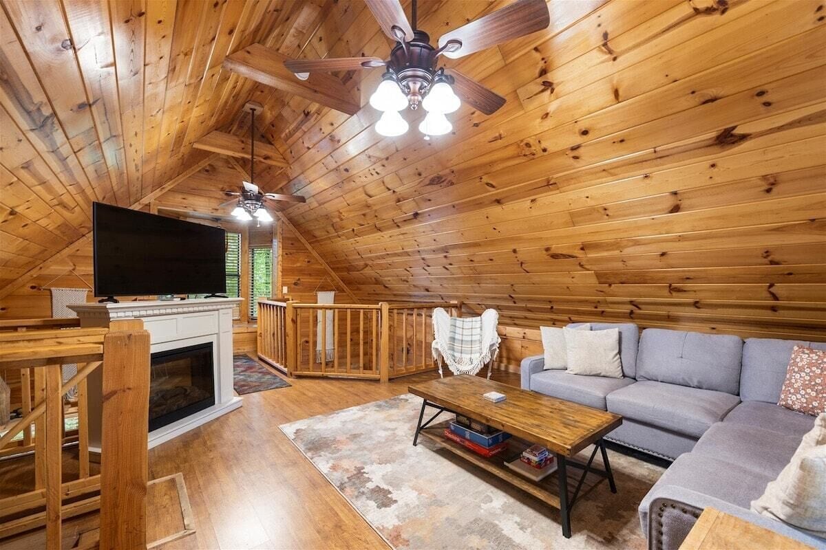 Un dormitorio acogedor en un ático con paredes de madera, ventilador de techo, televisor de pantalla plana, chimenea, sofá y mesa de comedor.