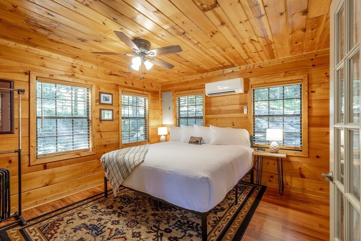 A wooden cabin bedroom with a bed, two bedside tables, a ceiling fan, and large windows.