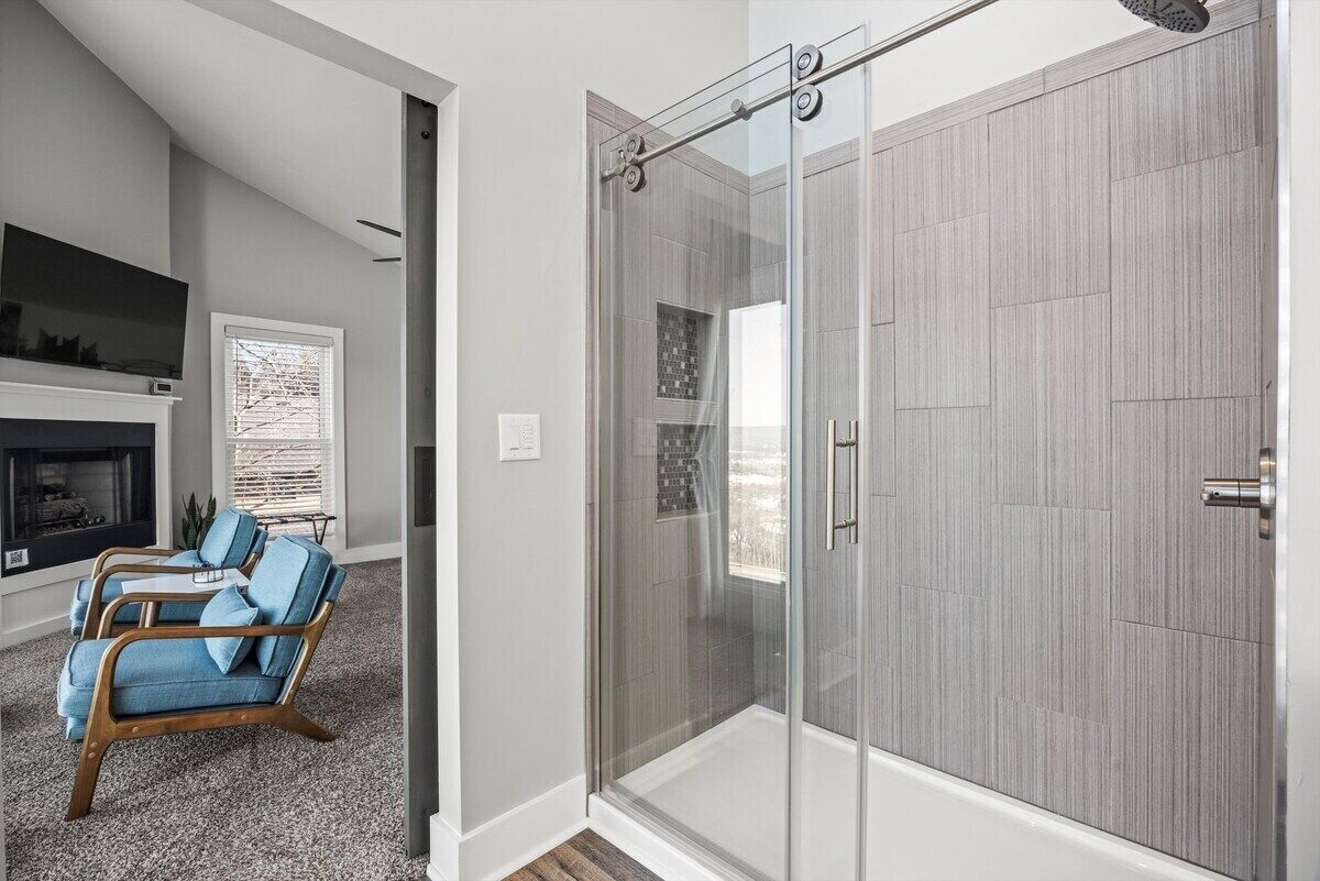Een moderne badkamer met een douchecabine van glas, een houten wastafelmeubel en een zithoek met een stoel en kussens.