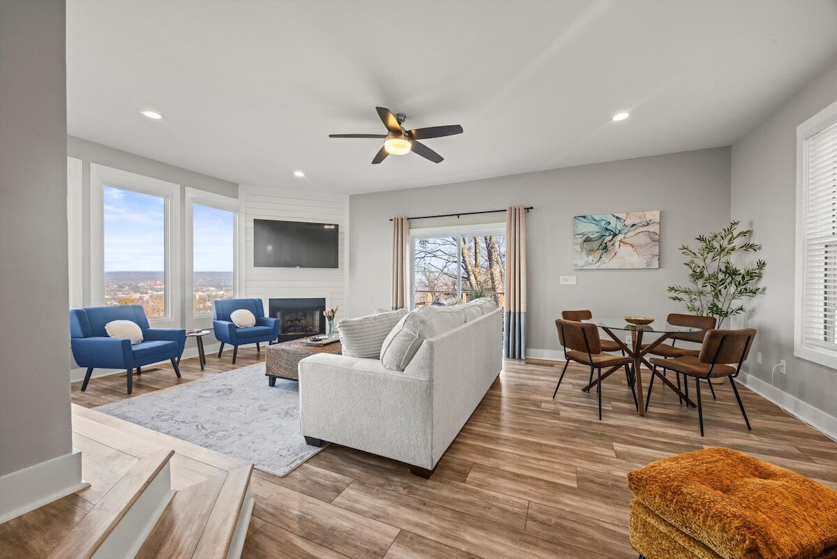 A spacious living room with a grey sofa, blue armchairs, a wooden floor, a ceiling fan, a fireplace, and a dining area with a table and chairs.