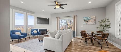A spacious living room with a grey sofa, blue armchairs, a wooden floor, a ceiling fan, a fireplace, and a dining area with a table and chairs.