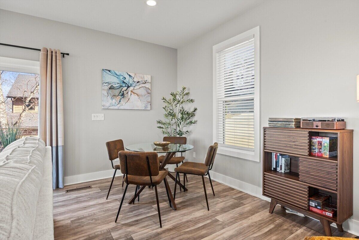 Uma área de refeições com mesa de vidro, cadeiras marrons, estante de livros e um quadro na parede.