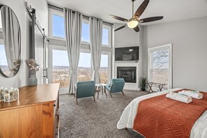 A hotel room with a bed, a desk, a chair, a small table, a ceiling fan, a fireplace, and large windows with a view. - Crestview Heights | Stunning City Views | Hot Tub (Chattanooga)