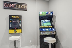 Ein Spielzimmer mit Spielautomaten, darunter Pac-Man und ein Golfspiel, und zwei weiße Barhocker.