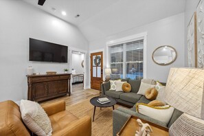 Ein Wohnzimmer mit einer Couch, einem Couchtisch und einem Fernseher. Es gibt ein Holzschränkchen und ein Fenster mit Jalousien.