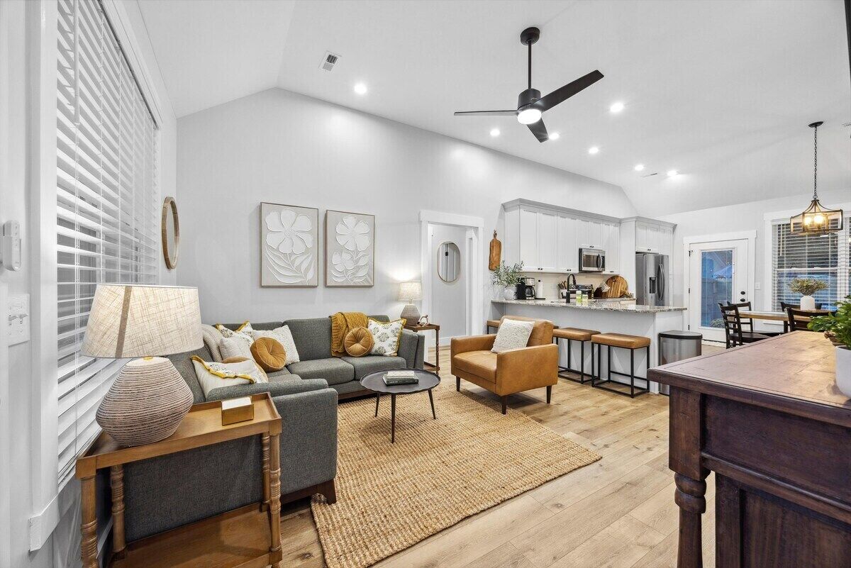 A modern living room with a sofa, armchairs, and a coffee table. A kitchen is visible in the background with a dining area.