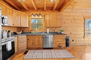 A wooden cabin-style kitchen with a stainless steel dishwasher, wooden cabinets, a granite countertop, and a window with a view of trees. - Crossbow Cabin | Lookout Mountain • Stunning Views (Rising Fawn)