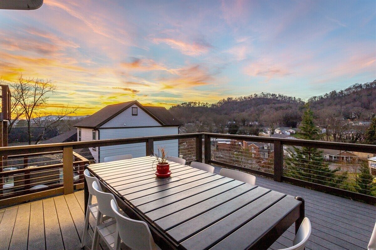 A wooden deck with a table and chairs, overlooking a residential area and hills during sunset.