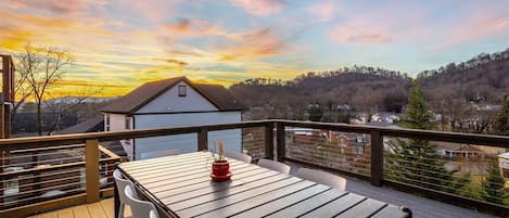 A wooden deck with a table and chairs, overlooking a residential area and hills during sunset.
