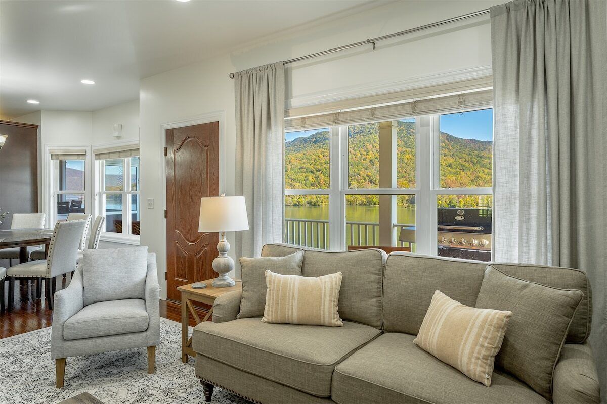 A living room with a sofa, armchair, and a table with a lamp. There is a window with a view of a forest and mountains.