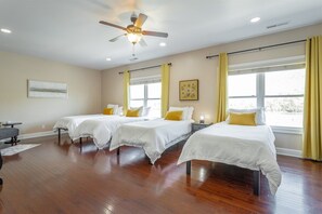 Quarto de hotel com três camas, um ventilador de teto e janelas amplas com cortinas.