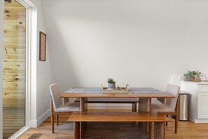 Un coin repas avec une table en bois, quatre chaises et un banc. On peut voir une petite plante sur la table et un tableau encadré au mur.