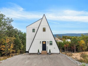 A white, triangular-shaped building with a black door, numbered 928, surrounded by trees and a clear sky. - Alpine A-Frames | 6 Luxe Cabins | Sleeps 60! (Rossville)