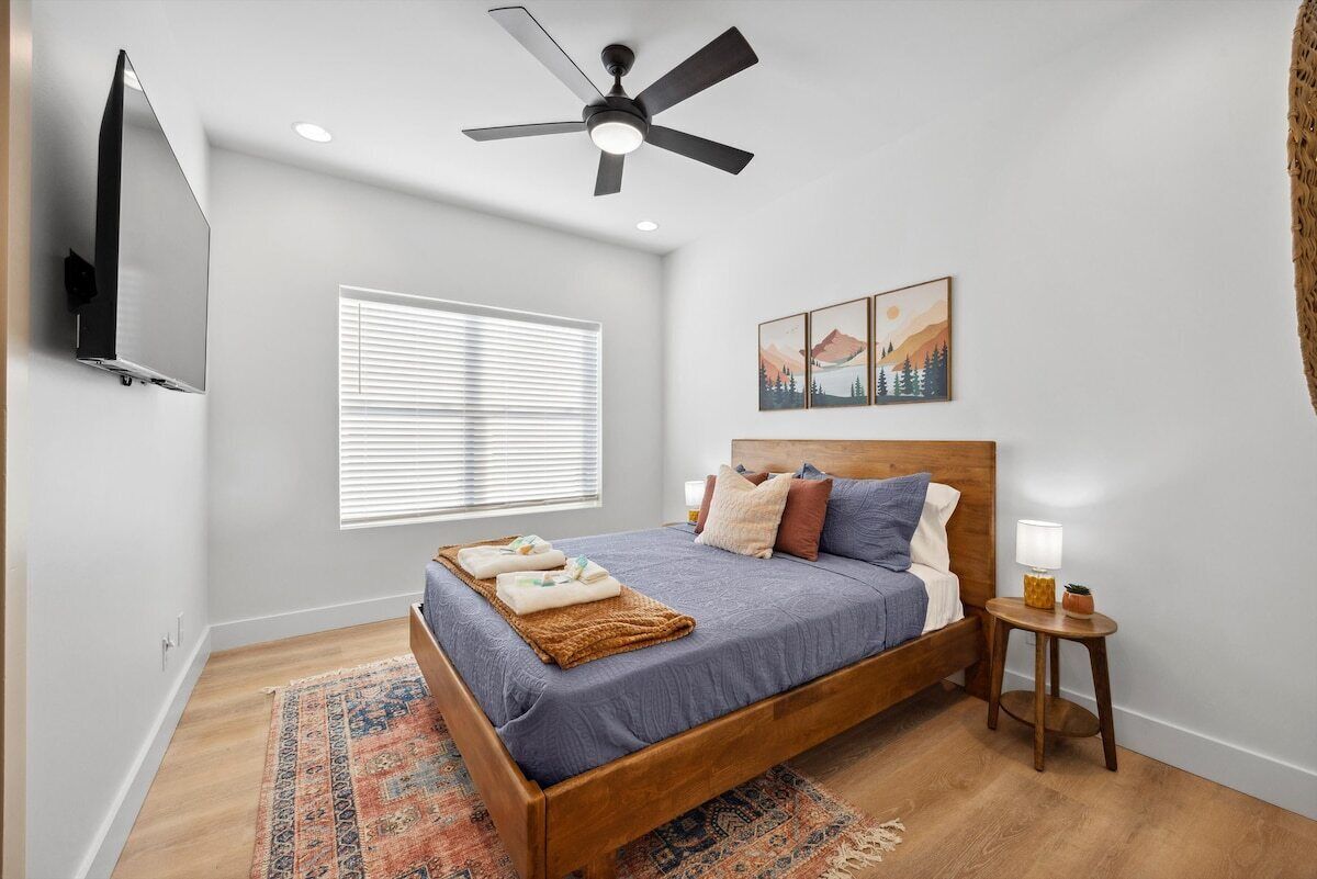 Ein Schlafzimmer mit einem Bett, einem Deckenventilator, einem Fernseher, einem Fenster mit Jalousien und Kunstwerken an der Wand.