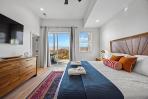 A modern bedroom with a large bed, a wooden dresser, a flat-screen TV, and a view of the outdoors. - Sunset Peak | A-Frame Bohemian Design | Hot Tub! (Rossville)