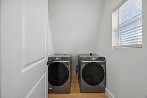 A laundry room with two front-loading washers and dryers, a window with blinds, and a wooden floor. - Sunset Peak | A-Frame Bohemian Design | Hot Tub! (Rossville)