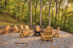 A forested area with a fire pit and wooden chairs arranged around it. - Bluebird Cottage | Near Downtown | Walking Trails! (Chattanooga)