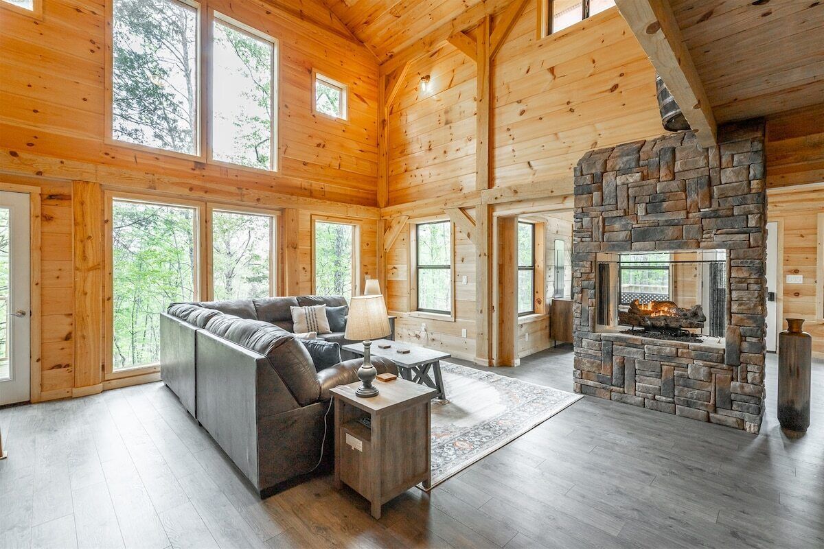 A log cabin interior with a stone fireplace, wooden walls, and large windows.