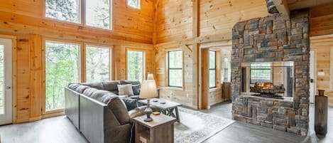 A log cabin interior with a stone fireplace, wooden walls, and large windows.