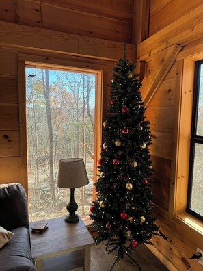 A cozy cabin interior with a Christmas tree, a lamp on a table, and a view of trees through a window.