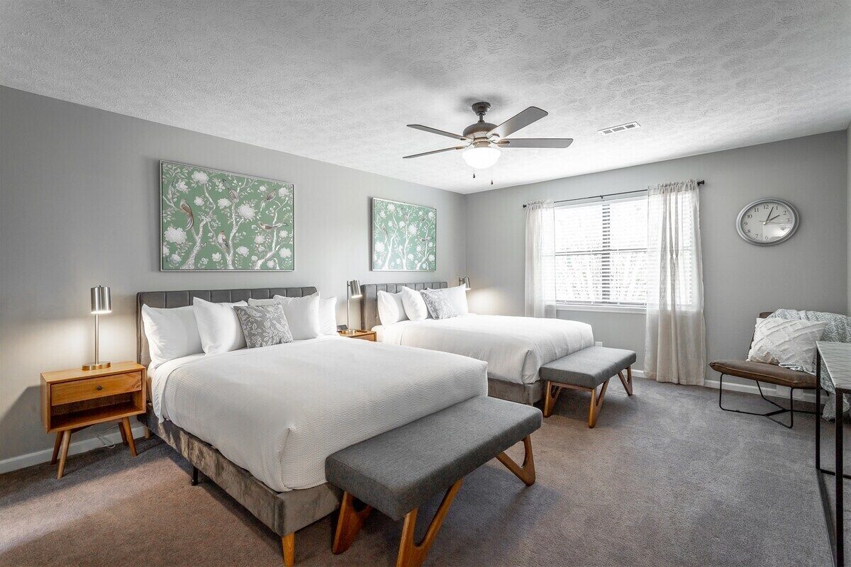 A bedroom with two beds, a ceiling fan, a bedside table with a lamp, a clock, and a window with curtains.