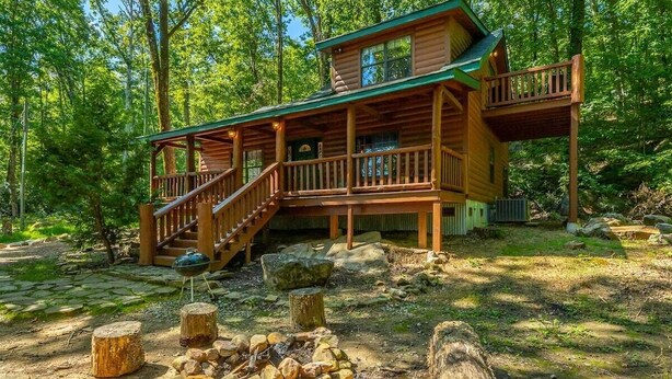 A wooden cabin with a deck, a staircase, and a fire pit in a forest setting. - Hideaway Lodge | Rustic W/ Hot Tub! Pet Friendly (Trenton)